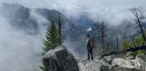 Persona equipada con mochila táctica y equipo de supervivencia, de pie en un paisaje desafiante con montañas y un río al fondo